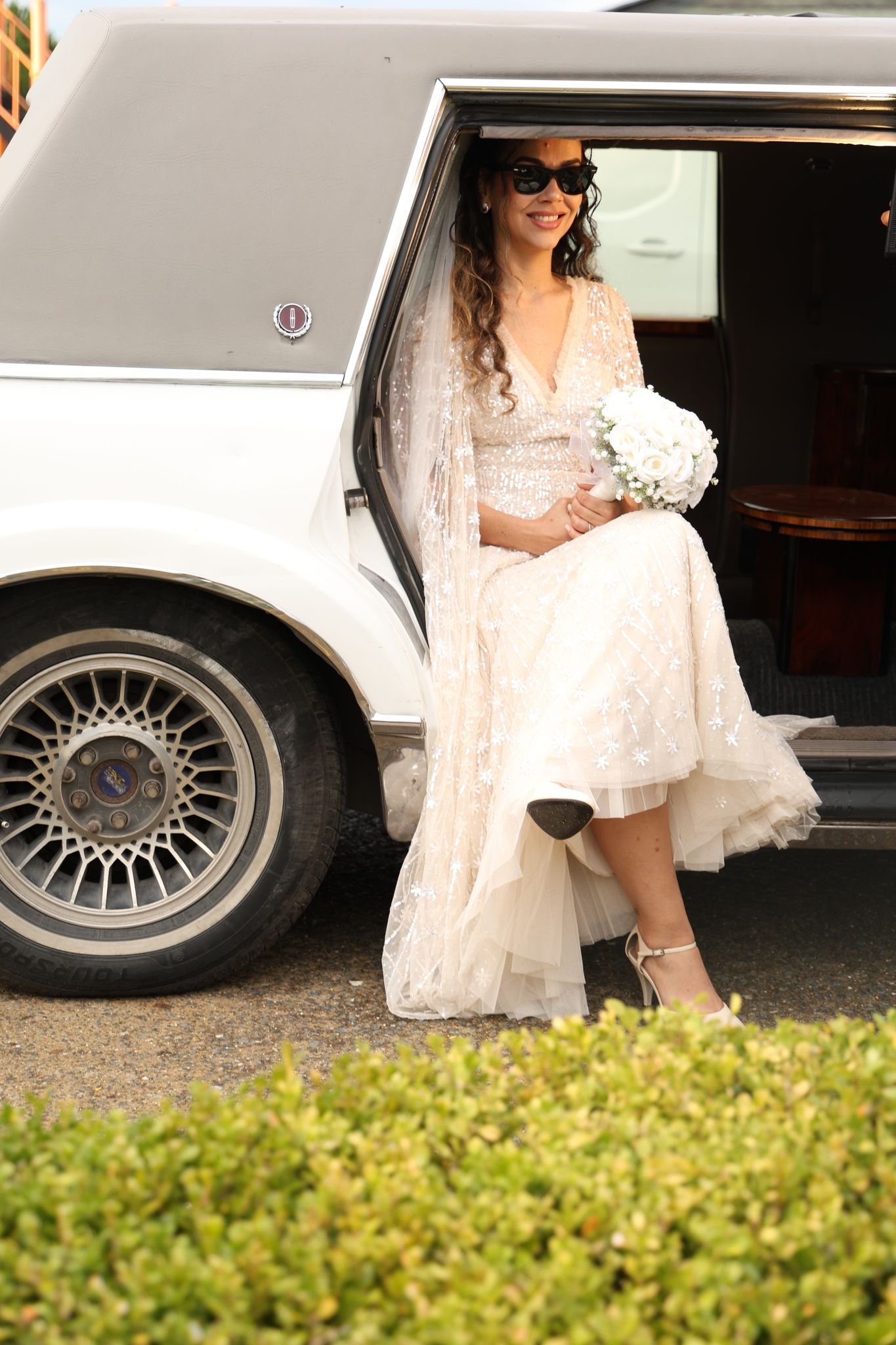 Bride in white dress stepping out of a limousine holding a bouquet, exuding elegance.